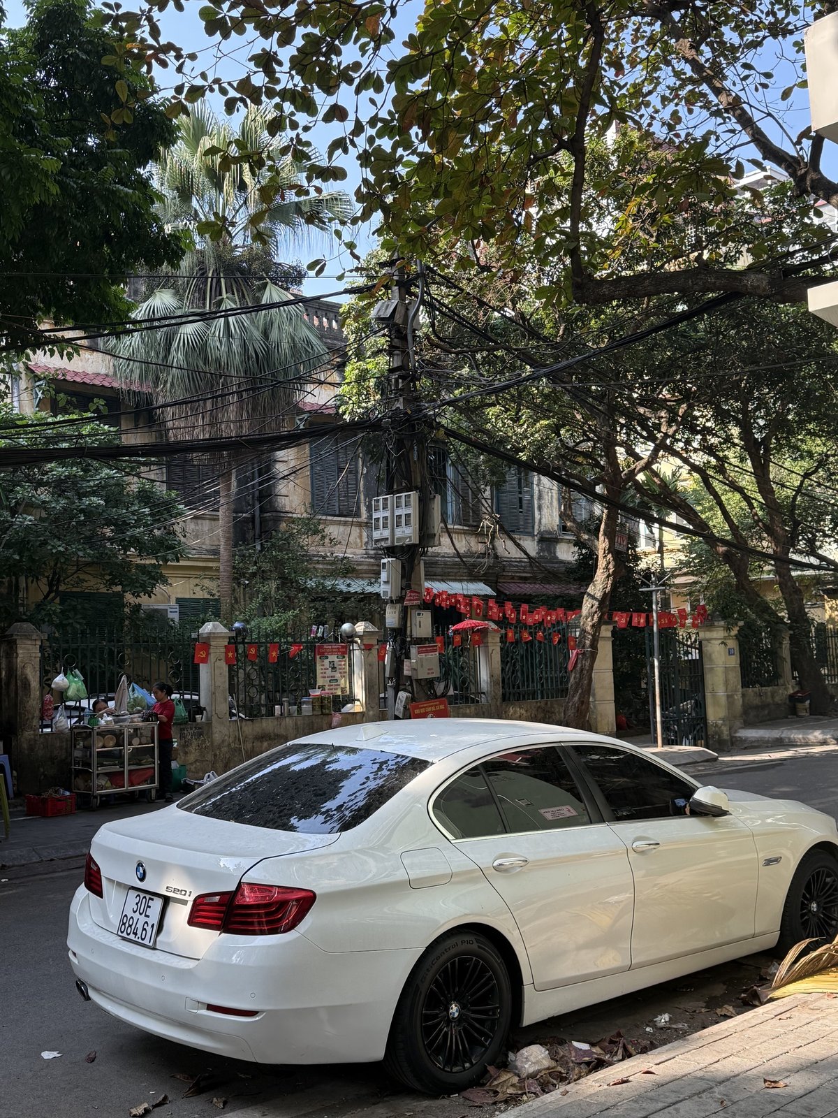 Hanoi side street with tangled overhead wires, apartment blocks, and parked traffic, illustrating everyday district texture
